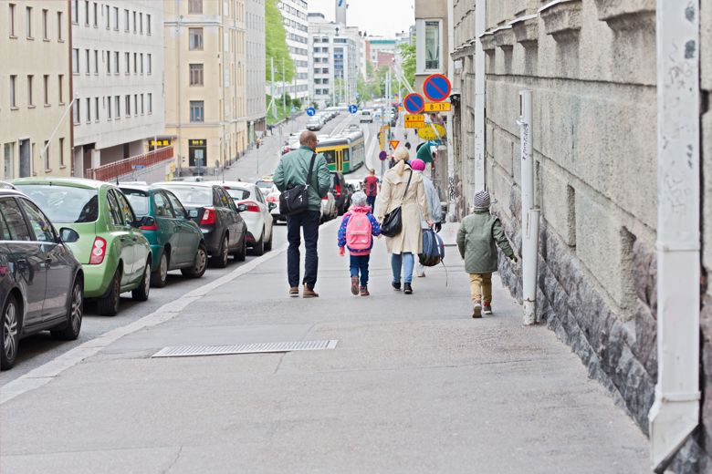 Alaspäin viettävä katu, jota ympäröi rakennukset. Kadun reunassa pysäköityjä autoja. Taustalla näkyy myös ratikka ja kadun reunassa muutama lehdessä oleva puu. Jalkakäytävällä selin kävelemässä aikuisia ja lapsia.