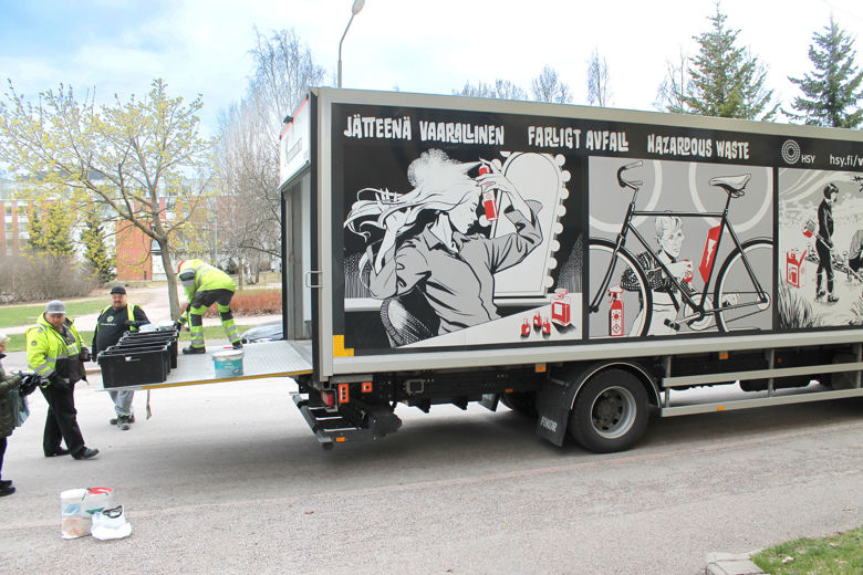 En lastbil som stannat på kanten av gatan.  Bilens bakgavellyft är nere och på den finns lådor och en person i varselkläder.