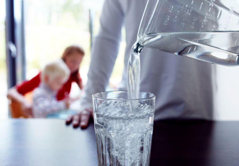 Mies kaataa vesikannusta vettä pöydällä olevaan lasiin. Taka-alalla nainen ja pieni lapsi istuvat pöydän ääressä..