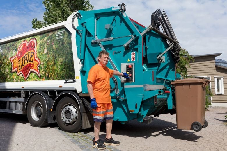 Biojäteastia tyhjennetään värikkääseen jäteautoon kuskin seisoessa sen edessä.