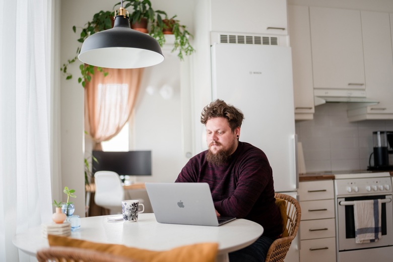 En man sitter i köket vid bordet med en bärbar dator framför sig.