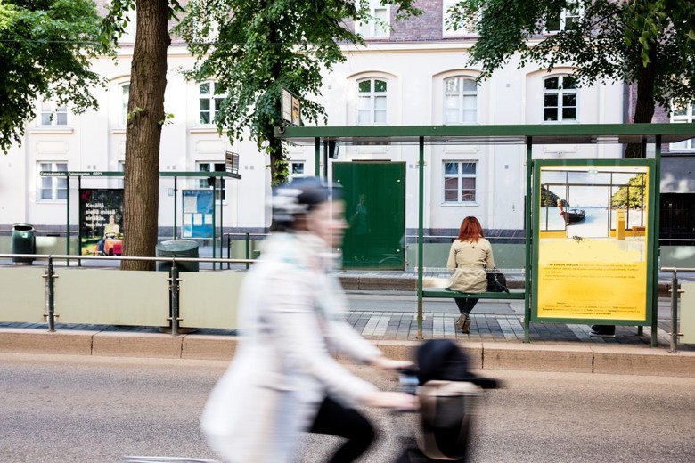 Polkypyöräilijä kiitää kadulla niin kovaa, että hän kuvassa hieman epätarkka. Pyöräilijan takana  ratikkapysäkki, jonka penkillä istuu ihminen.  Pysäkin molemmin puolin puita. Taustalla rakennusrivistö.