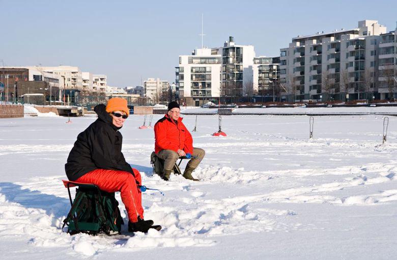 Two ice fishers in the foreground with the city behind them on a sunny day. The ice is covered by a layer of snow.