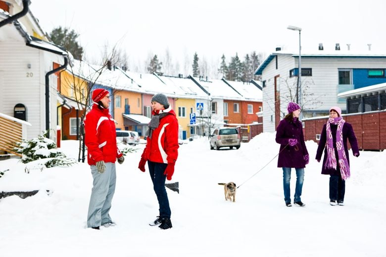 People walking and talking on a snowy residential street. A dog on a leash. In the background, there are parked cars and two-story buildings connected to each other in a winding formation.