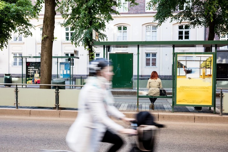 En cyklist kommer så fort längs gatan att bilden är lite otydlig. Bakom cyklisten är en spårvagnshållplats där en person sitter på bänken.  På båda sidorna om hållplatsen står träd. I bakgrunden syns en rad byggnader.
