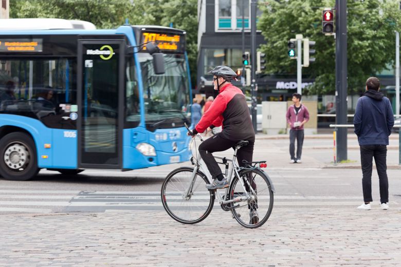 Linja-auto ajaa suojatien ohi, jossa pyöräilijä ja jalankulkijat odottavat liikennevalon vaihtumista vihreäksi.