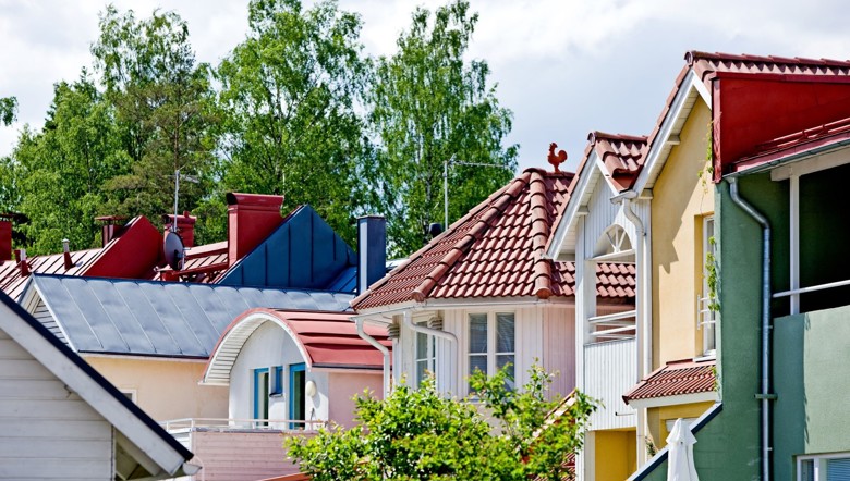 Erivärisiä ja -muotoisia taloja on vierekkäin. Taloista näkyvät vain yläosat, joten huomio kiinnittyy kattojen hauskoihin muotoihin. Erään talon katolla on kukon muotoinen tuuliviiri. Taustalla on puita,